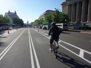 Lone bicyclist on Pennsylvania Avenue bike lane in early morning; buses queued in background